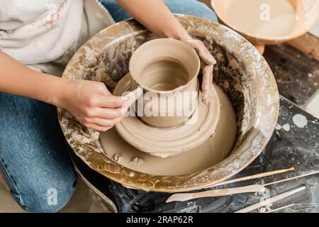 Draufsicht einer jungen Töpferin in Schürze, die die Form einer Tonvase mit Holzwerkzeug auf einem drehenden Töpferwerkzeug in Keramikwerkstätten, Tonformen und für herstellt Stockfoto