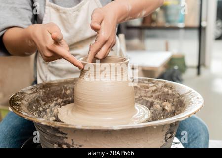 Zugeschnittener Blick auf eine junge Kunsthandwerkerin in einer Schürze, die Vase aus nassem Ton herstellt und mit drehenden Töpferrädern in einer unscharfen Keramikwerkstatt arbeitet, Töpferei C. Stockfoto