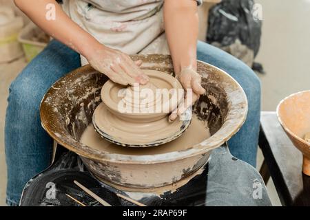 Freigestellte Ansicht einer jungen Künstlerin in Schürze, die Ton formt und während der Arbeit an Töpferrädern in der Nähe von Holzwerkzeugen im Kunststudio, geschickter Topf Stockfoto