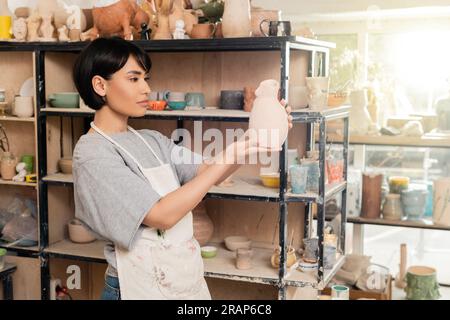 Junge braune asiatische Kunsthandwerkerin in Schürze, die Tonskulptur in der Nähe eines Regals mit Regalen in einer Werkstatt für verschwommene Keramik im Hintergrund bei Sonnenuntergang hält, Kunsthandwerk Stockfoto