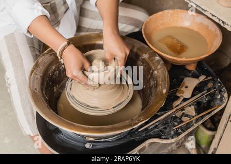 Draufsicht einer jungen Kunsthandwerkerin, die nassen Lehm auf einem Töpferrad in der Nähe von Werkzeugen und Schüssel mit Wasser in einer Keramikwerkstatt, einem Arbeitsplatz im Töpferstudio und cr formt Stockfoto