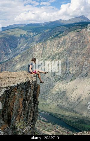 Gefährliche Fotos in der Natur. Eine Frau sitzt am Rand einer steilen Klippe über einer hohen Klippe und baumelt mit ihren Beinen. Tödliches Risiko für Fotografie. Sommer-Alta Stockfoto