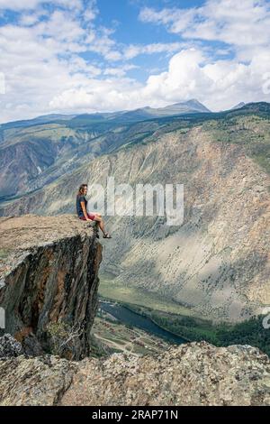 Die Frau sitzt am Rand einer steilen Klippe über einer tiefen Klippe, deren Beine nach unten hängen. Chulyshman-Tal, Katu-Yaryk-Pass, Altai. Fahren Sie rund um die W Stockfoto