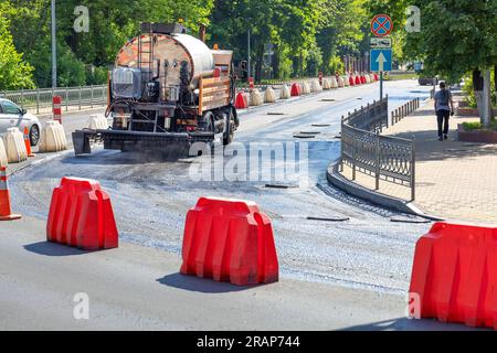 Ein Lkw mit einem großen Tankwagen spritzt flüssiges Bitumen auf eine Baustelle mit Kunststoffabsperrung auf einer Stadtstraße, bevor er an einem sonnigen Sommertag asphaltiert wird. Stockfoto