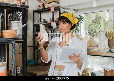 Junge brünette asiatische Kunsthandwerkerin in Kopftuch und Arbeitskleidung hält Tonskulptur und digitales Tablet während der Arbeit in einer Keramikwerkstatt, kreative PR Stockfoto