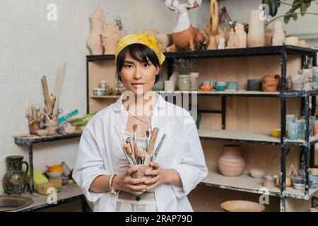 Junge brünette asiatische Kunsthandwerkerin in Arbeitskleidung und Kopftuch, die Töpferwerkzeuge hält und in der Keramikwerkstatt vor die Kamera schaut, kreativ Stockfoto