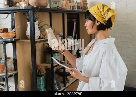 Seitenansicht einer jungen asiatischen Kunsthandwerkerin in Kopftuch und Arbeitskleidung mit digitalem Tablet mit leerem Bildschirm und Tonskulptur und keramischem Stand Stockfoto