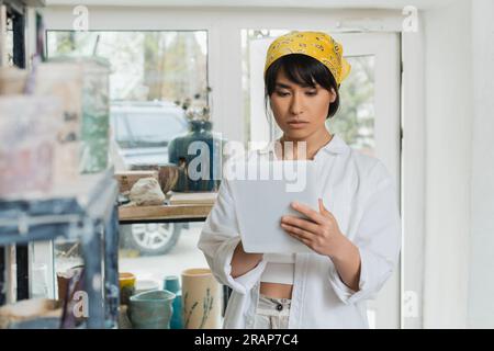 Porträt einer jungen braunen asiatischen Kunsthandwerkerin in Kopftuch und Arbeitskleidung mit einem digitalen Tablet im Hintergrund in einer unscharfen Keramikwerkstatt Stockfoto