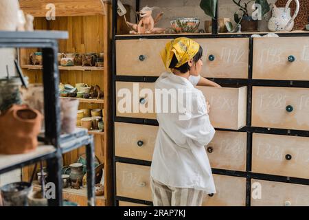 Seitenansicht einer jungen braunen asiatischen Kunsthandwerkerin im Kopftuch und Arbeitskleidung, die den Schrank öffnet, während sie im Keramikstudio steht und arbeitet, Keramik-Worksho Stockfoto