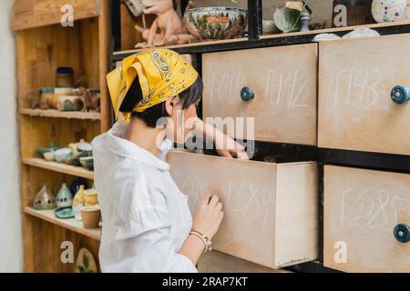 Seitenansicht einer jungen asiatischen Künstlerin mit Kopftuch und Arbeitskleidung, die den Schrank öffnet, während sie in einer verschwommenen Töpferwerkstatt, einer Töpferwerkstatt mit Ski arbeitet Stockfoto