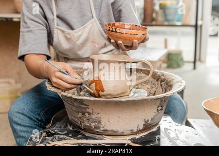 Schnittbild einer jungen Künstlerin bei Schürzenmalerei auf Tonkanne auf Töpferrad neben Holzwerkzeugen n Tisch in einer unscharfen Keramikwerkstatt, Tongestaltung Stockfoto