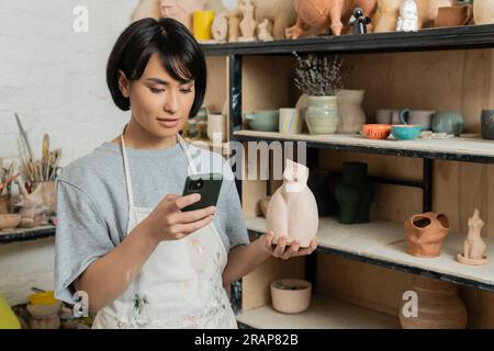 Junge braune asiatische Kunsthandwerkerin in einer Schürze mit Smartphone und Keramikskulptur in der Hand, während sie in der Nähe eines Regals mit Produkten in einer Keramikwerkstatt stand Stockfoto