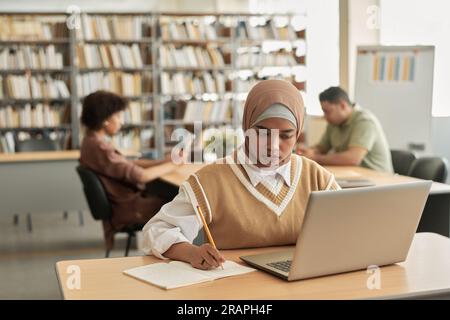 Muslimischer Student, der Laptop im Studium benutzt, während er an einem Tisch in der Bibliothek sitzt Stockfoto