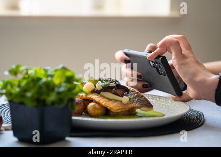 Fotografieren Sie mit dem Smartphone gerösteten Branzino mit marinierten Pilzen und Butterkartoffeln. Veröffentlichen und Teilen von Lebensmittelbildern in sozialen Medien. Stockfoto