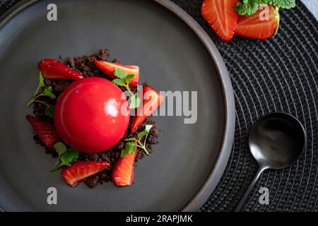 Draufsicht auf einen kugelförmigen Quark mit Erdbeeren und Brownies. Rotes Dessert mit glatten Oberflächen und Spiegelglasur auf dem schwarzen Teller mit schwarzem Löffel. Stockfoto