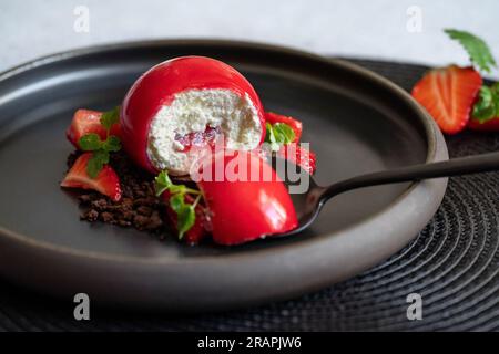 Selektiver Fokus eines kugelförmigen Quark mit Erdbeersoße auf einem schwarzen Teller. Ein Kinderspiel auf dem schwarzen Löffel. Erdbeeren und Brownies. Roter Nachtisch. Stockfoto