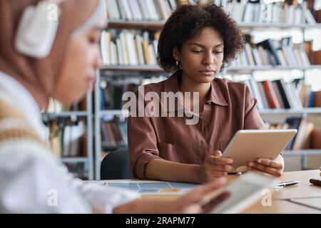 Afroamerikanischer Student, der sich den Online-Unterricht auf einem Tablet-pc ansieht, während er mit anderen Schülern in der Bibliothek am Tisch lernt Stockfoto