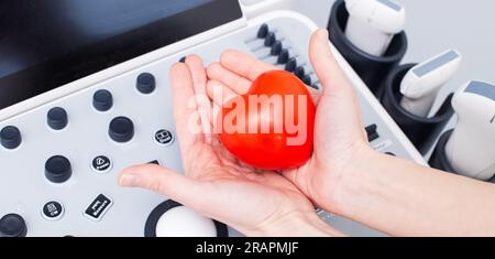 Hände eines Ultraschallarztes, der ein rotes Herz vor Ultraschalldiagnosegeräte hält. Gesundheitsvorsorge und Medizinisches Konzept. B Stockfoto