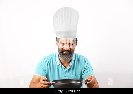 Porträt eines bärtigen indischen Chefkochs mit T-Shirt und Kochmütze. Fröhliche, lächelnde Ausdrücke, weißer Hintergrund. Den Pfannenwender und den Frittierlöffel in der Hand halten Stockfoto