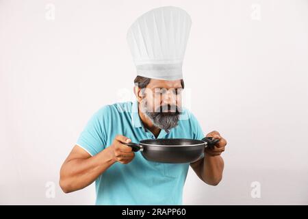 Porträt eines bärtigen indischen Chefkochs mit T-Shirt und Kochmütze. Fröhliche, lächelnde Ausdrücke, weißer Hintergrund. Den Pfannenwender und den Frittierlöffel in der Hand halten Stockfoto