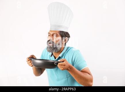 Porträt eines bärtigen indischen Chefkochs mit T-Shirt und Kochmütze. Fröhliche, lächelnde Ausdrücke, weißer Hintergrund. Den Pfannenwender und den Frittierlöffel in der Hand halten Stockfoto