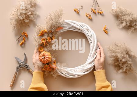 Frau, die einen Herbstkranz mit natürlichen trockenen Blumen und Kürbis auf einem beigen Tisch macht. Draufsicht. Thanksgiving-Dekoration. Stockfoto