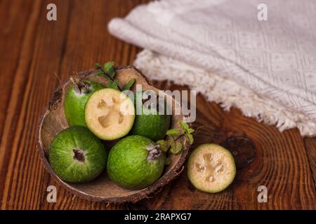 Grüne Feijoa in Kokosnussschalen mit Löffel auf Holzhintergrund. Reife tropische Früchte, Minzblätter, Ananasguava, rohe vegane Speisen. Kalorienarm, Stockfoto