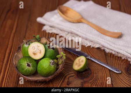 Grüne Feijoa in Kokosnussschalen mit Löffel auf Holzhintergrund. Reife tropische Früchte, Minzblätter, Ananasguava, rohe vegane Speisen. Kalorienarm, Stockfoto