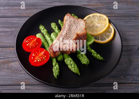 Gegrillte Ersatzrippchen mit grünem Spargel, Tomaten und Zitrone auf einem Teller auf grauem Holzhintergrund Stockfoto