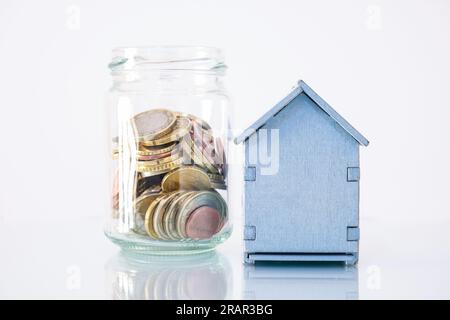 Ein kleines Holzhaus mit einem Glasbehälter daneben, in dem sich Euro-Münzen ansammeln, das Konzept der Sparsamkeit, die Effizienz im Haushalt. Stockfoto