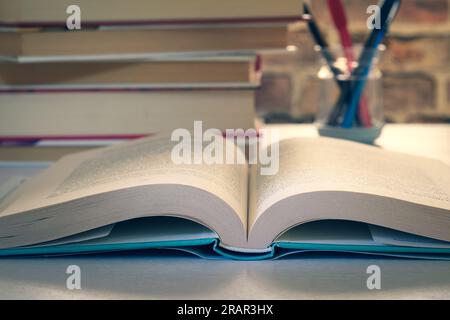 Offenes festes Buch auf einem Schreibtisch, gestapelte Bücher und ein Stifthalter im Hintergrund, Lernkonzept, Forschung, Wissen. Stockfoto