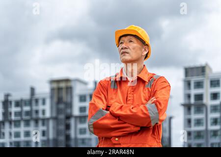 Seriöser und entschlossener asiatischer Ingenieur mittleren Alters in orangefarbenem Overall mit Schutzhelm, der seine Arme kreuzte, während er stolz vor seinem Multi-Stori stand Stockfoto