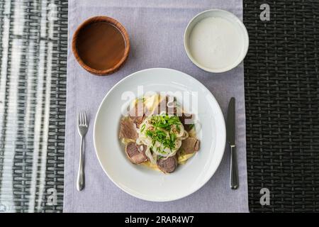 Nahaufnahme des Schüssels Beshbarmak auf einem Teller auf dem Tisch. Große Stücke gedünstetes Fleisch und Zwiebeln. Beshbarmak – Kasachisches Nationalgericht, zubereitet mit Fleisch und Pasta Stockfoto