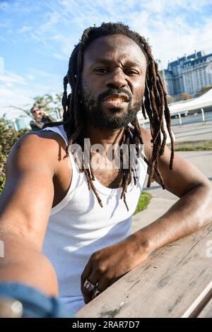 Vertikales Selfie-Porträt eines jungen Afrikaners mit Dreadlocks und Bart, der draußen bei Sonnenuntergang sitzt, mit einem wütenden Gesicht in die Kamera schaut, nimmt Stockfoto