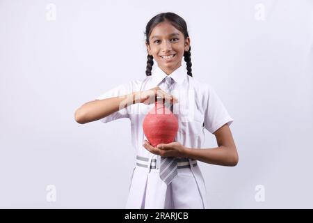 Ein glückliches indisches Schulmädchen, das ein Sparschwein in der Hand hält, zeigt fröhlich verschiedene Stimmungen von Sparen und Bankkonzepten Stockfoto