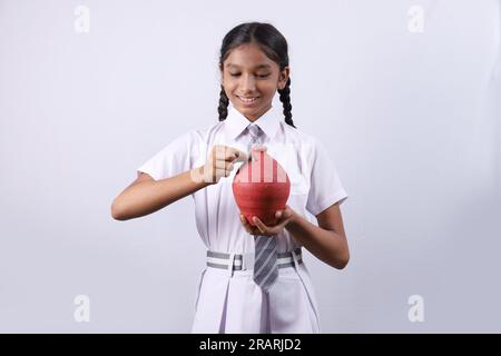 Ein glückliches indisches Schulmädchen, das ein Sparschwein in der Hand hält, zeigt fröhlich verschiedene Stimmungen von Sparen und Bankkonzepten Stockfoto