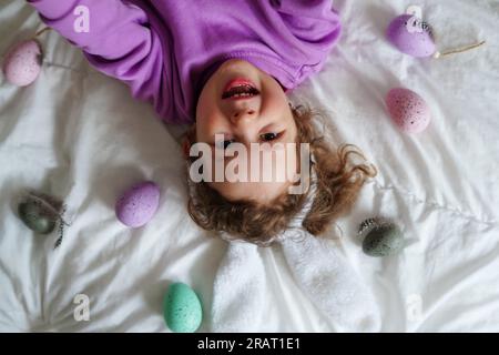Frohe Ostern. Süßes, wunderschönes Mädchen mit lockigen Haaren und Hasenohren, die auf dem Bett liegen, mit bunten ostereiern. Lifestyle-Foto Stockfoto