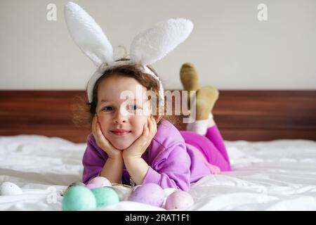 Frohe Ostern. Süßes, wunderschönes Mädchen mit lockigen Haaren und Hasenohren, die auf dem Bett liegen, mit bunten ostereiern. Lifestyle-Foto Stockfoto