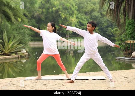 Junge Kinder, die früh am Morgen Yoga-Übungen in einem grünen öffentlichen Park machen. Wir können Wasserkörper sehen. Kinder sind gesundheitsbewusst. Internationaler Yoga-Tag. Stockfoto