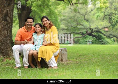 Glückliche indische Familie mit einem Mädchen, das zusammen auf dem Gras sitzt und ihre Familienzeit am frühen Morgen genießt. Die Familie schaut in die Kamera. Stockfoto