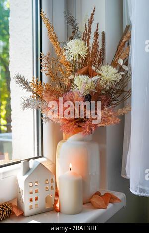 Süßes Zuhause. Stille Lebensdetails im Haus auf einem Holzfenster. Herbstdekor am Fenster, getrocknete Blumen, Kerzen und Spielzeughaus Stockfoto