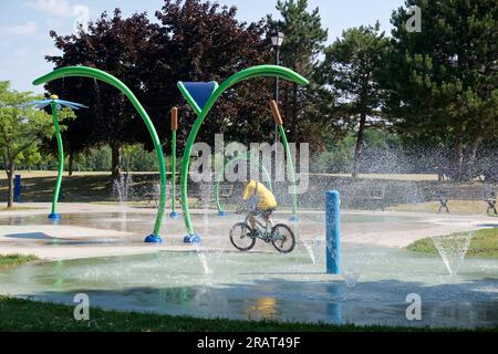 Toronto, Ontario/Kanada - 07/22/2022: Junge, der zwischen den Sprinklern mit dem Fahrrad fährt. Ein Junge kühlt sich an einem heißen Sommertag mit Wasser ab Stockfoto