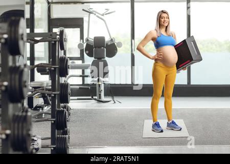 Porträt einer schwangeren Frau in Sportbekleidung, die eine Stufe-Aerobic-Plattform im Fitnessstudio hält Stockfoto