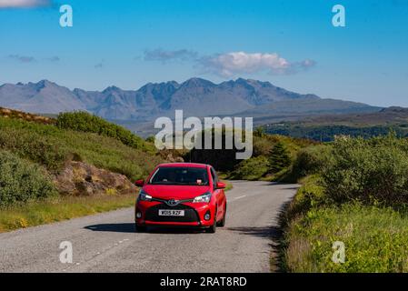 Isle of Skye, Schottland, Vereinigtes Königreich. 5. Juni 2023 Kleines rotes Auto und malerischer Blick auf die Cuillin Hills mit blauem Himmel. Isle of Skye, Vereinigtes Königreich. Stockfoto