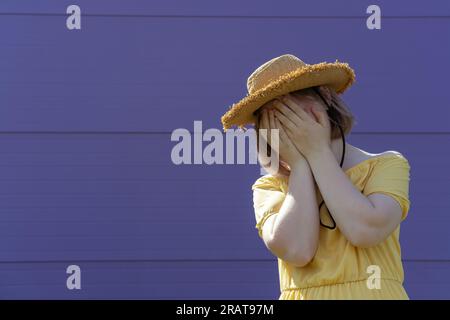 Eine frustrierte asiatische Teenagerin mit Hut und Kleid bedeckt ihr Gesicht mit ihren Händen auf einem lila Hintergrund. Psychisches Gesundheitskonzept. Stockfoto