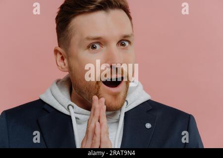 Verblüffter Mann in Sweatshirt und schwarzer Jacke, offener Mund, zusammengepresste Handflächen, Ungläubigkeit, isoliert auf pinkfarbenem Hintergrund Stockfoto