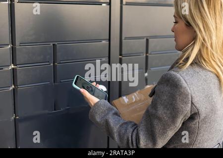 Schöne Frau, die ein Paket aus einem intelligenten elektronischen Paketschließfach aus Stahl abholt, automatische Briefkästen. Paczkomat-Lieferservice, Abholung Stockfoto