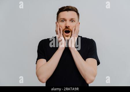 Ein bärtiger Mann mit Insektenaugen im schwarzen T-Shirt, Hände auf den Wangen. Reaktion auf etwas erstaunliches. Isoliert auf blauem Hintergrund. OMG-Konzept. Stockfoto