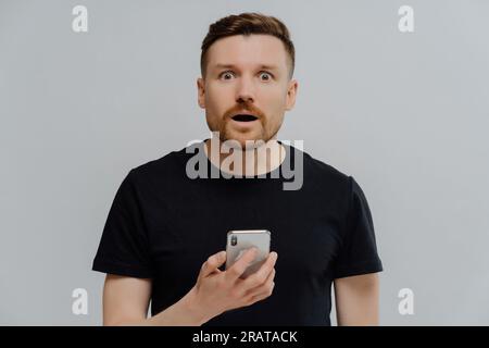 Ein überraschter Mann mit Bart im schwarzen T-Shirt, der ein Handy in der Hand hat. Auf schockierende Nachrichten reagieren. Innenhaltung, Online-Kommunikation. Stockfoto