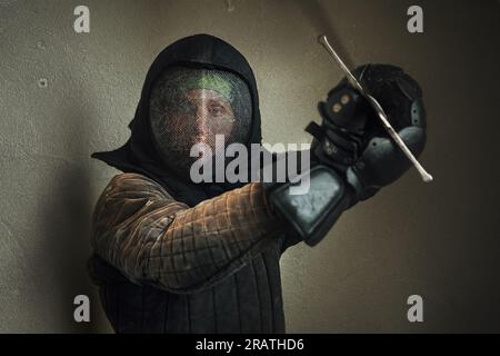 Porträt eines Mannes in schwarzer historischer Zäunenpanzerung und Schutzhelmmaske mit einem langen mittelalterlichen scharfen Schwert, das für aggressiv vorbereitet wurde Stockfoto